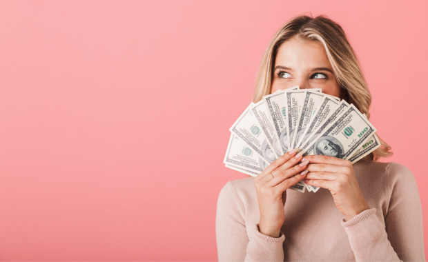 Woman holding a fan of money against a pink background – illustrating the cost of a hen do.
